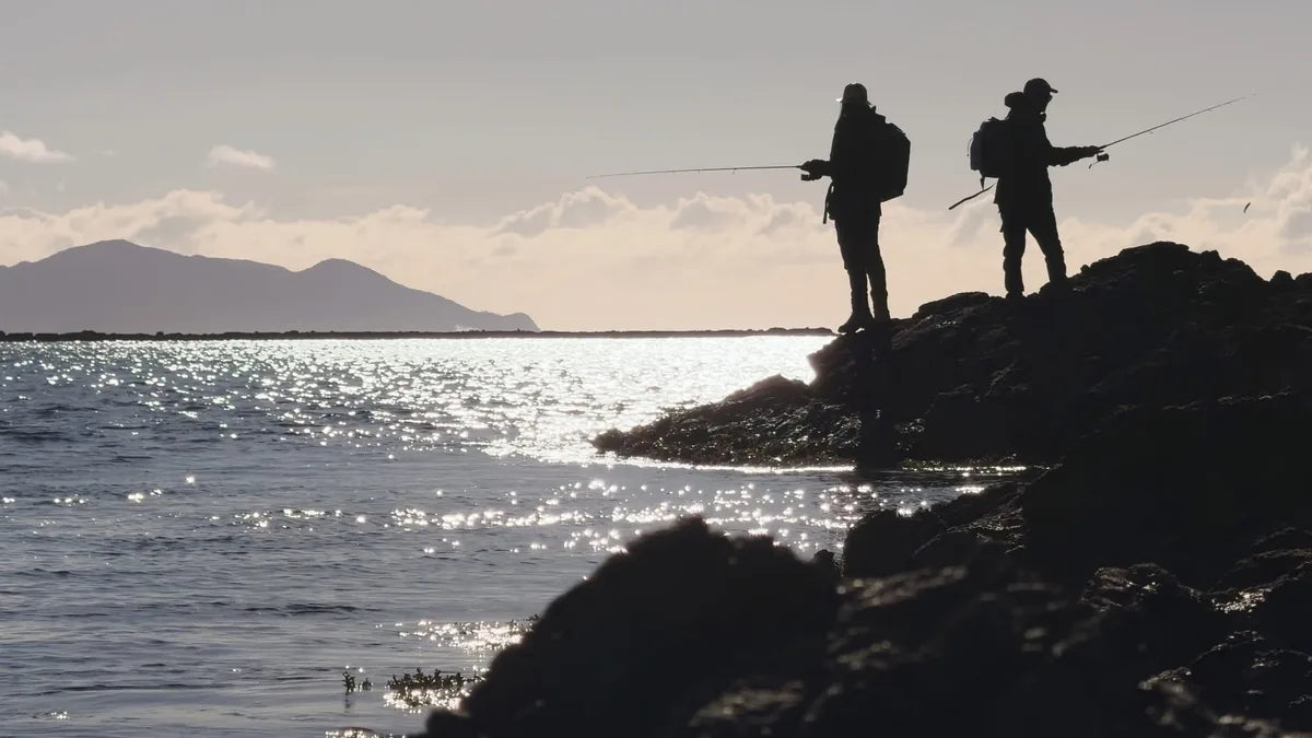 Two anglers silhouetted on coastal rocks at sunset, casting over a sparkling sea